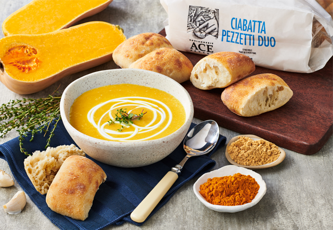 Butternut squash soup with ACE® Ciabatta Pezzetti Rolls on a dark blue cloth surrounded by squash, seasoning and a spoon